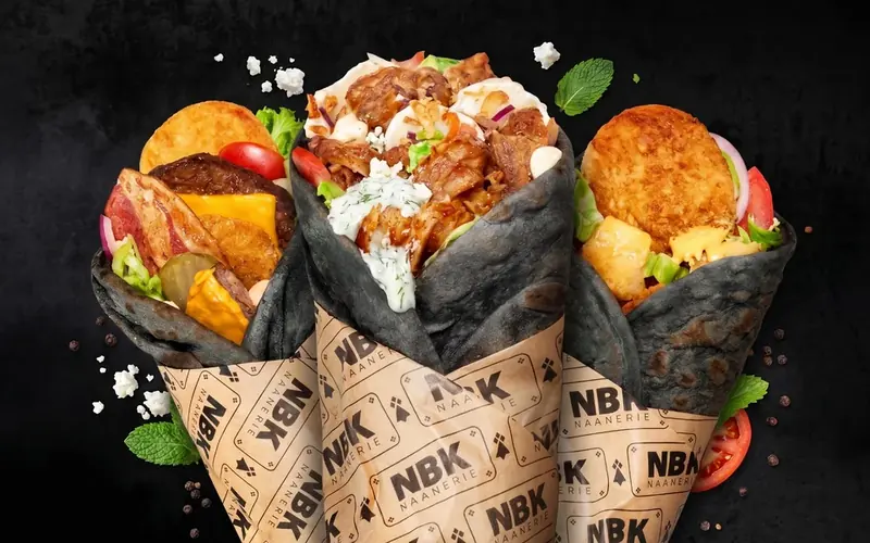 Trois Black Naans NBK au pain noir, dans leur papier siglé, sur fond noir : steak, bacon et cheddar, kebab de poulet sauce blanche, et une galette croustillante