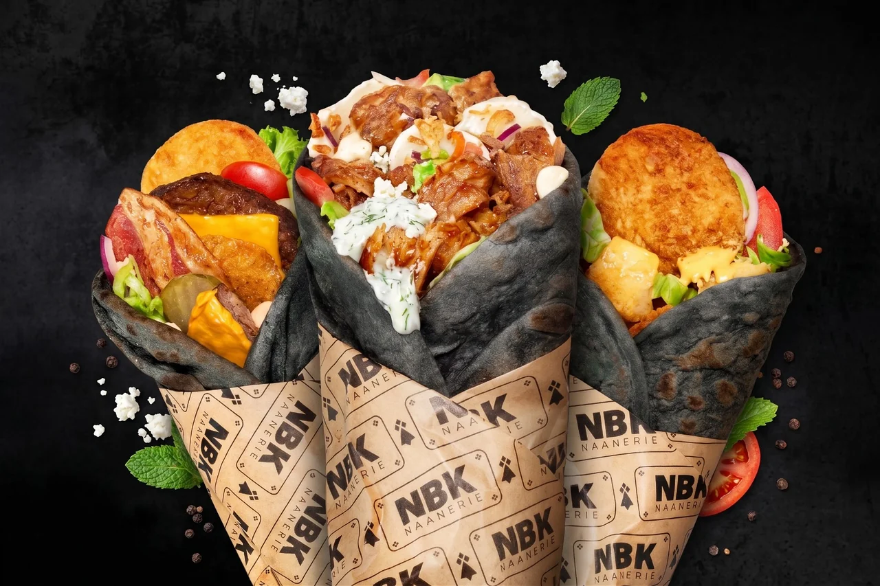 Trois Black Naans NBK au pain noir, dans leur papier siglé, sur fond noir : steak, bacon et cheddar, kebab de poulet sauce blanche, et une galette croustillante