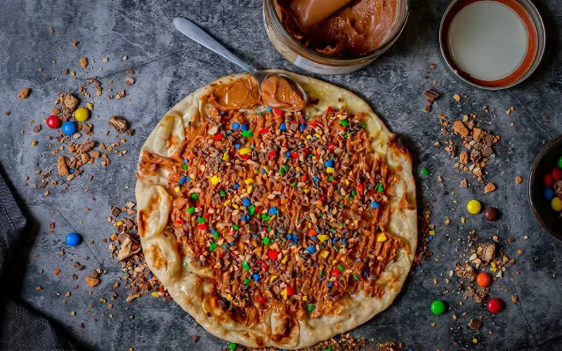 Un sweet naan NBK vu de dessus, nappé de pâte à tartiner et couvert d’éclats de biscuits et de bonbons chocolatés colorés, à côté du pot entamé, sur une table en pierre grise