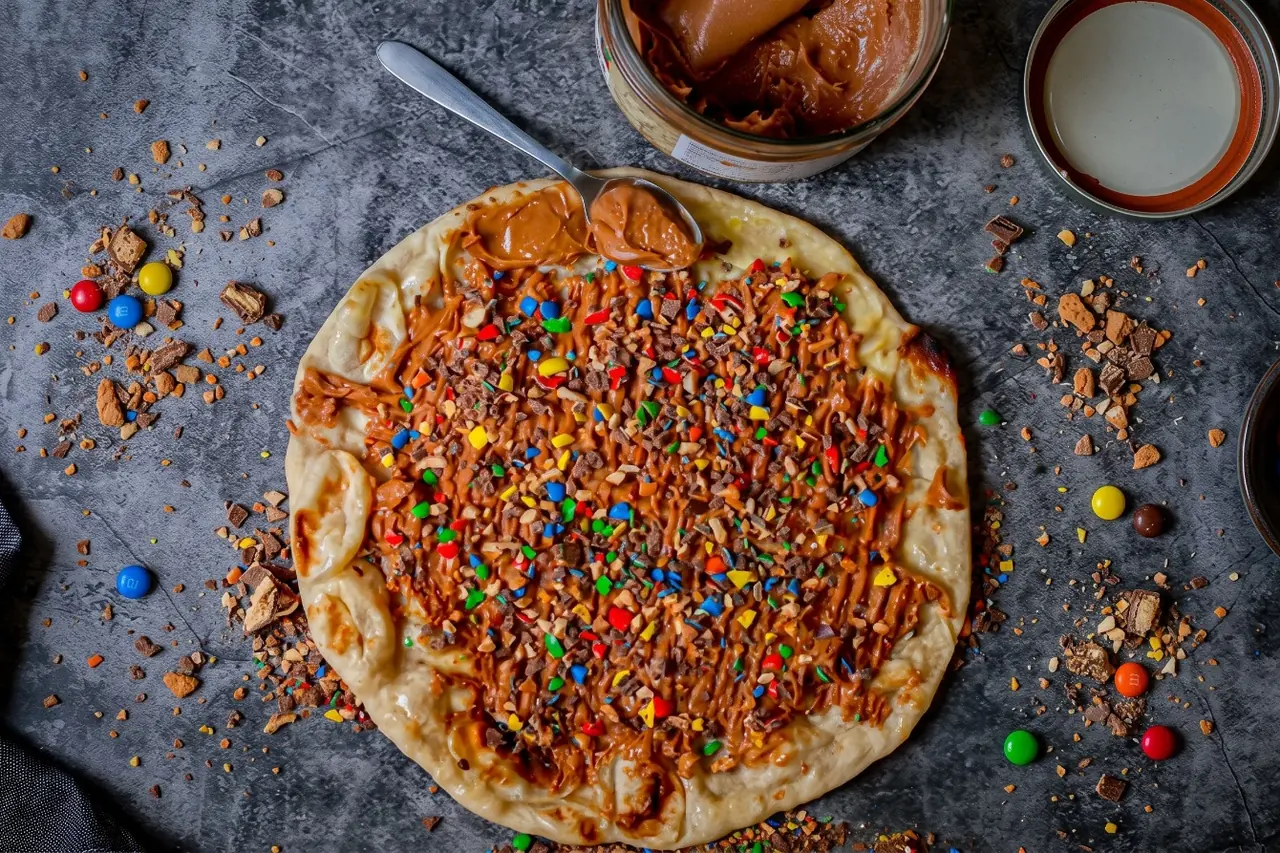 Un sweet naan NBK vu de dessus, nappé de pâte à tartiner et couvert d’éclats de biscuits et de bonbons chocolatés colorés, à côté du pot entamé, sur une table en pierre grise
