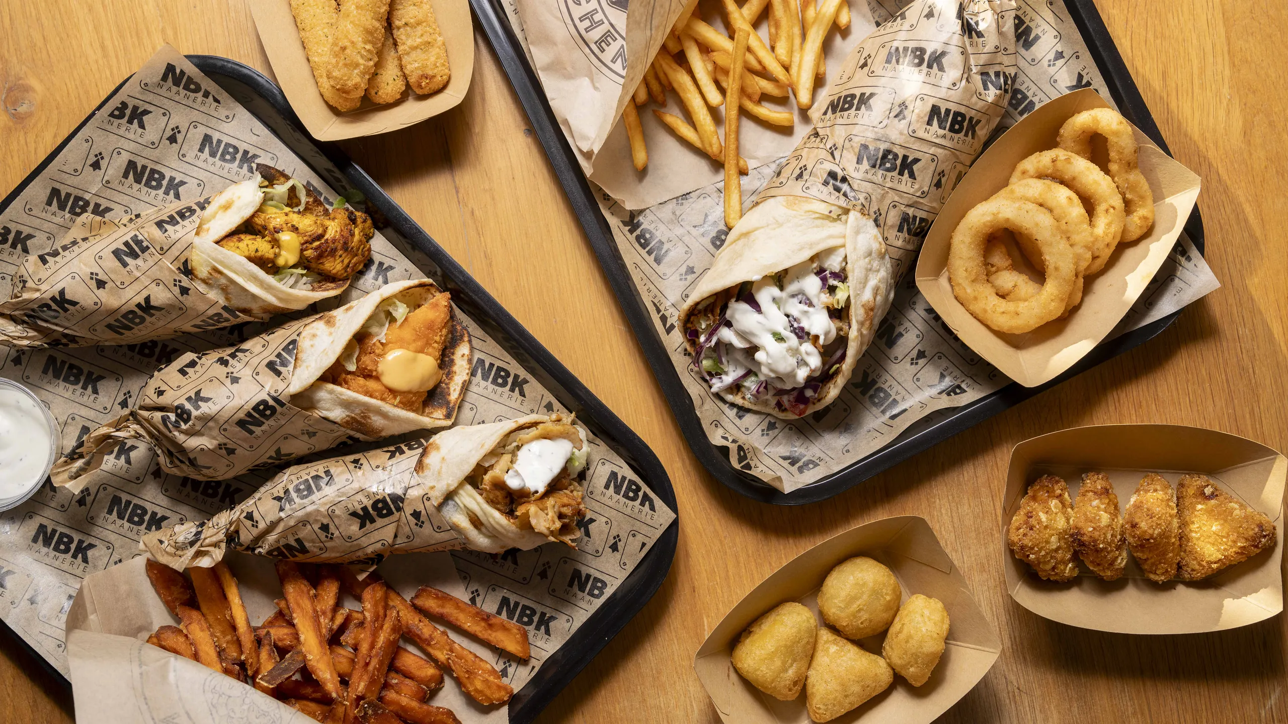 Deux plateaux NBK vus de dessus : cheese naans dans leur papier siglé, frites de patate douce, onion rings et nuggets