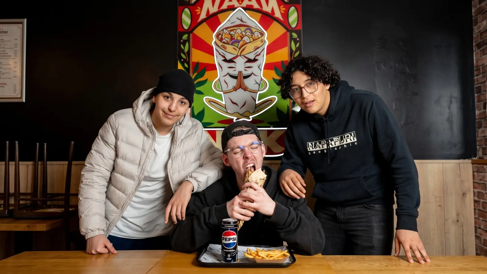 Trois clients NBK autour d’une table, l’un mordant dans un naan garni, devant l’affiche illustrée « Le Naan » et un plateau de frites
