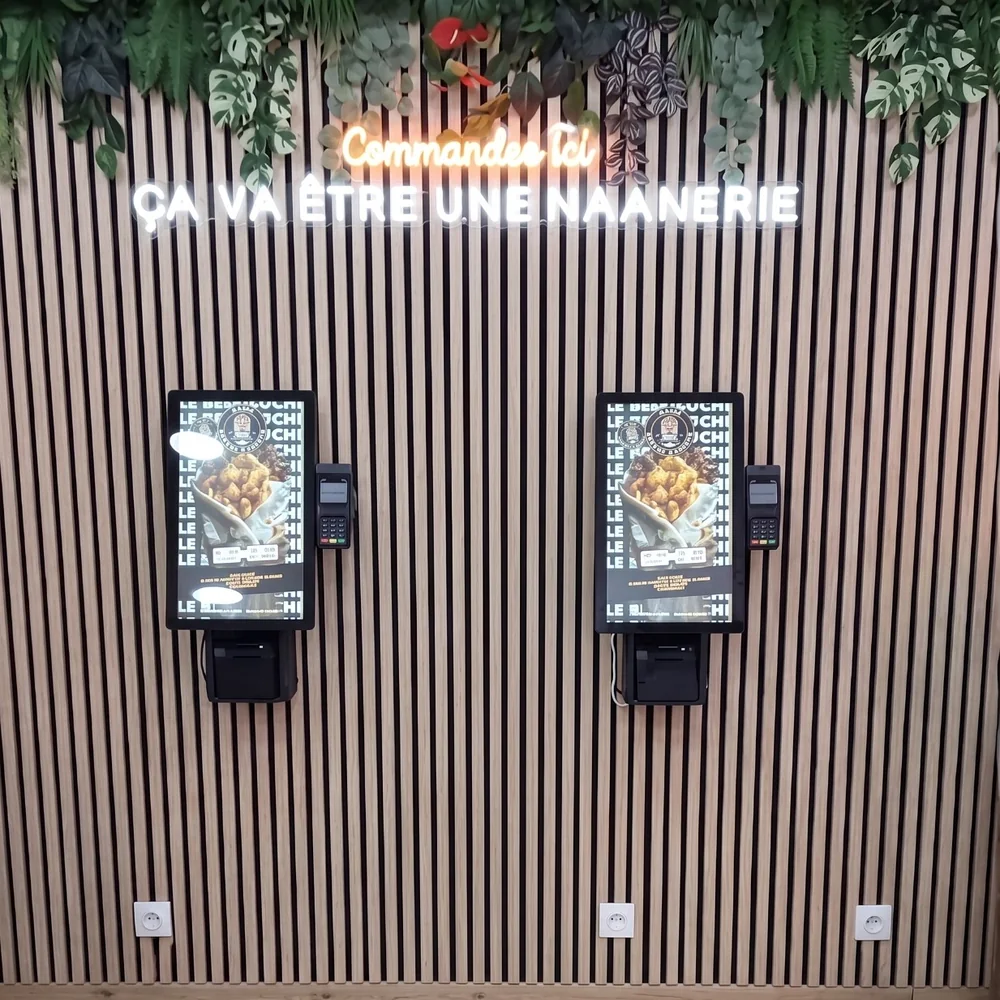 Deux bornes de commande NBK sur un mur de lattes de bois, sous les néons « Commandez ici » et « Ça va être une Naanerie »