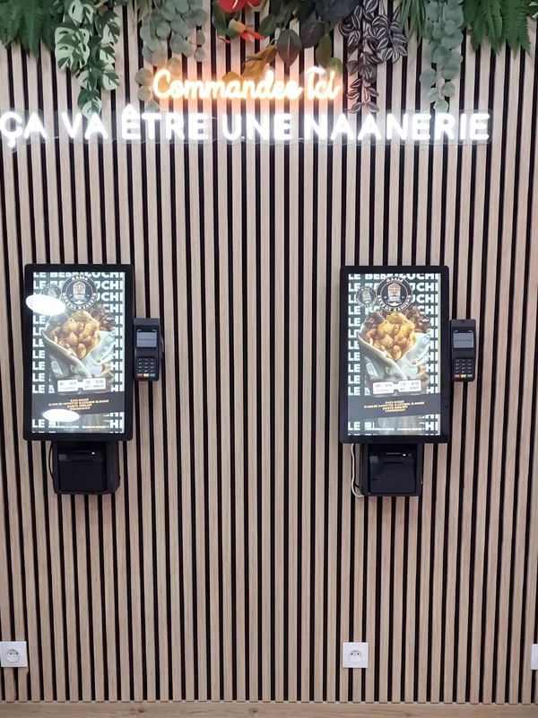 Deux bornes de commande NBK sur un mur de lattes de bois, sous les néons « Commandez ici » et « Ça va être une Naanerie »